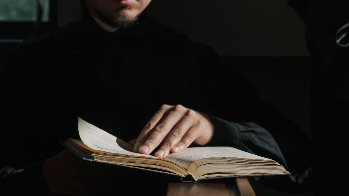 A man in dark clothing reads a Bible in a dimly lit church, highlighting faith and reflection.