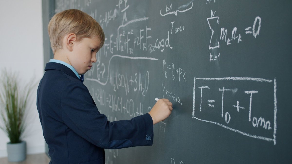 A boy in a suit writing complex math equations on a classroom blackboard.