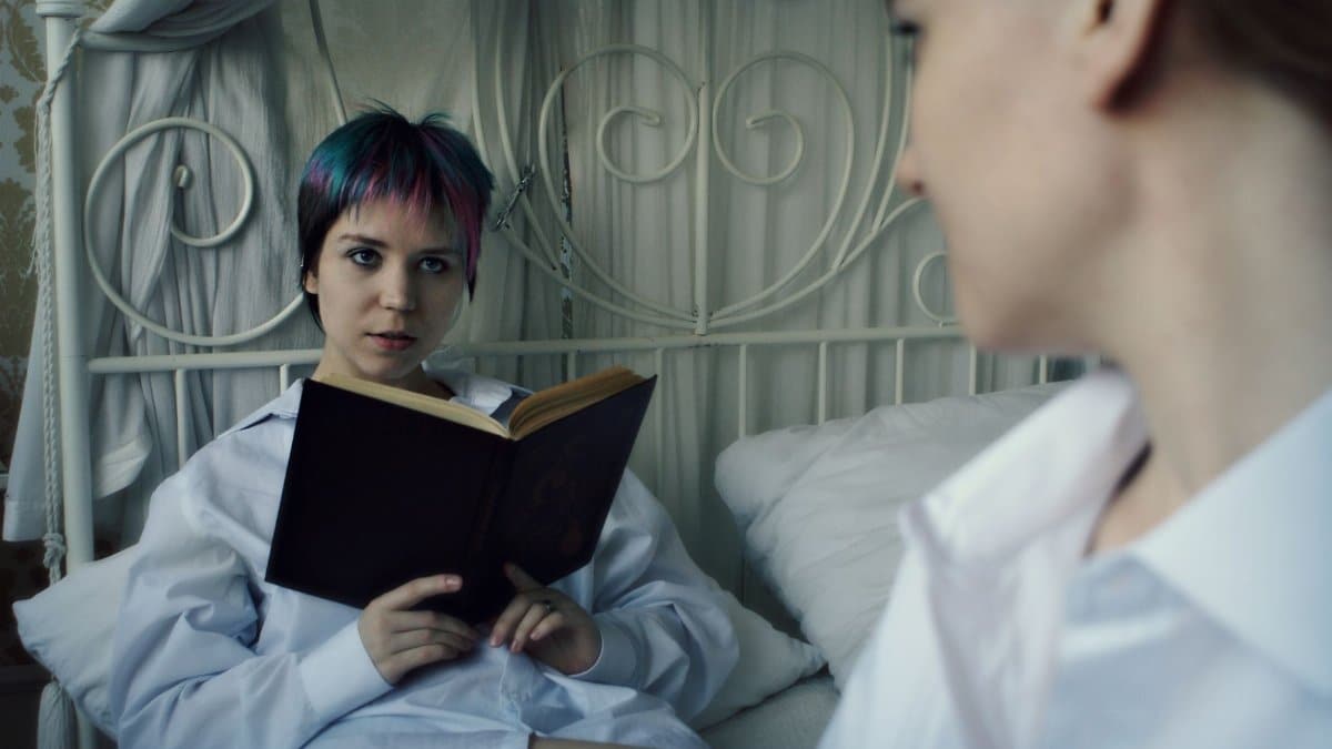 Serious young female with short multicoloured hair looking at friend while chilling on bed and reading book