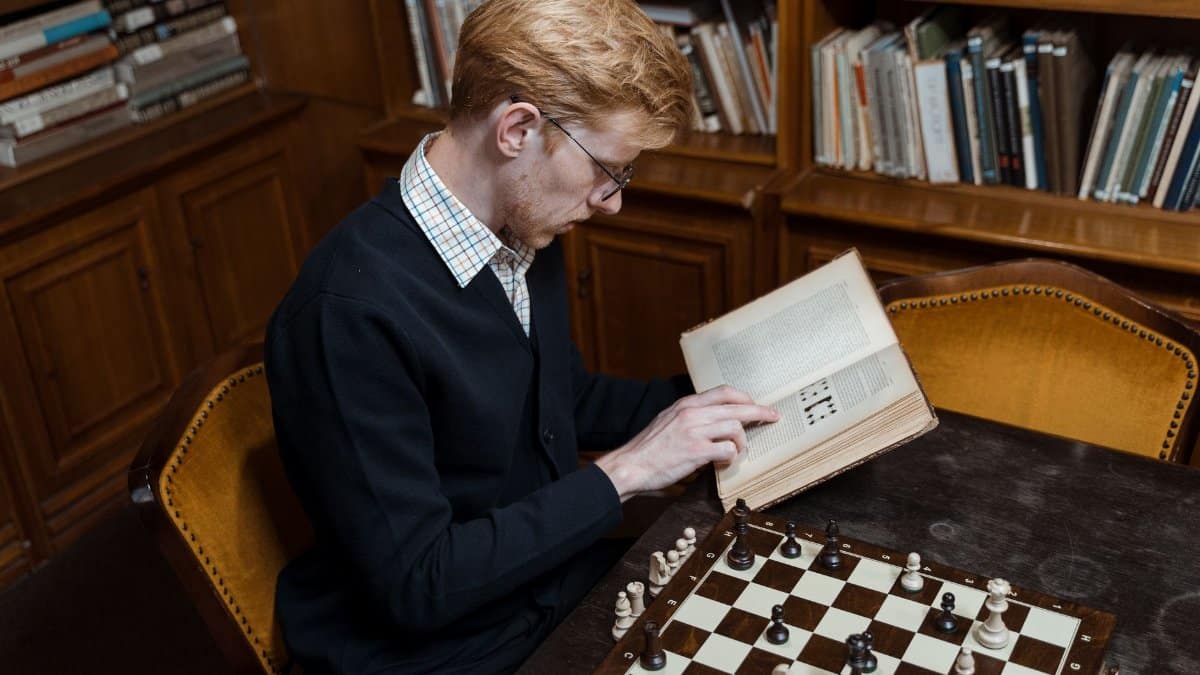 Focused man reading and playing chess in a cozy library setting.