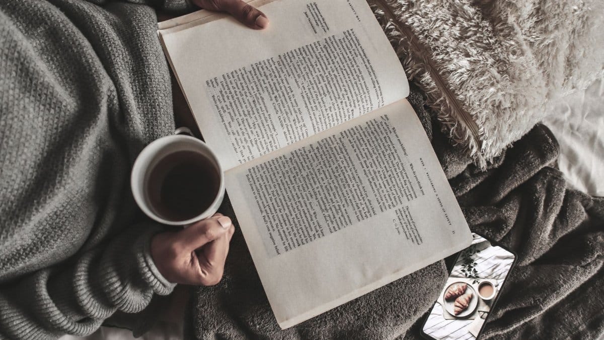 From above of crop anonymous female with mug of hot tea reading book under plaid near smartphone at home