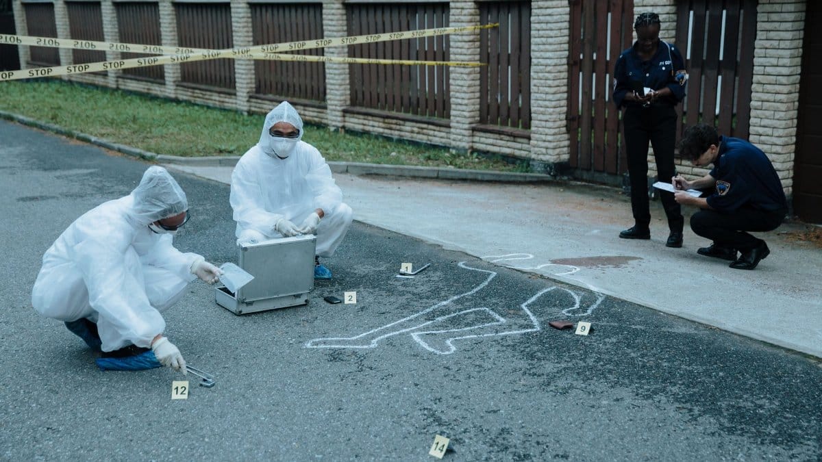 Police officers and investigators examine a crime scene with evidence markers and chalk outline.