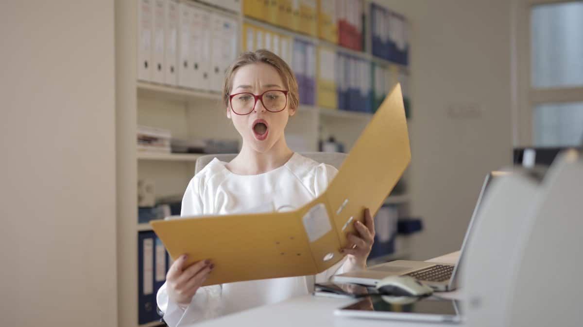 Surprised woman with eyeglasses reading a file in an office filled with shelves of records.