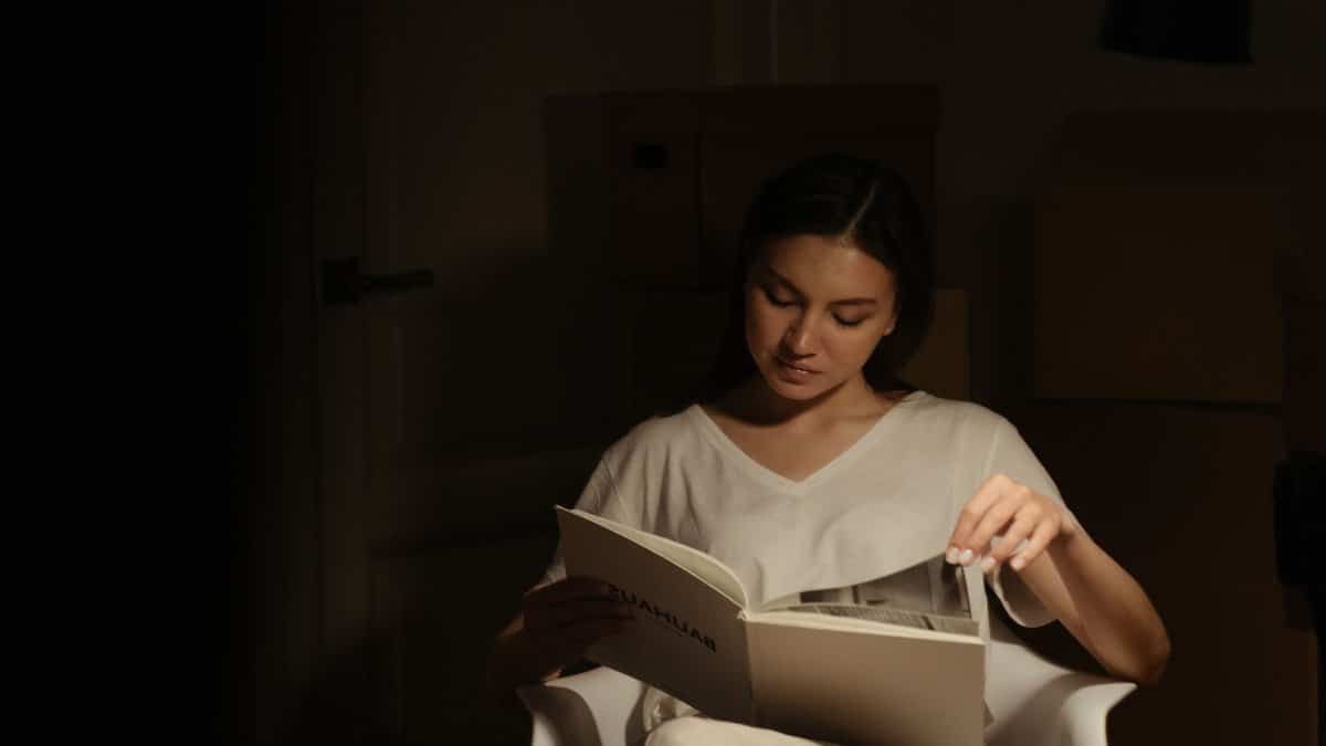 A woman sits in a dim room reading a book, creating a serene ambiance.