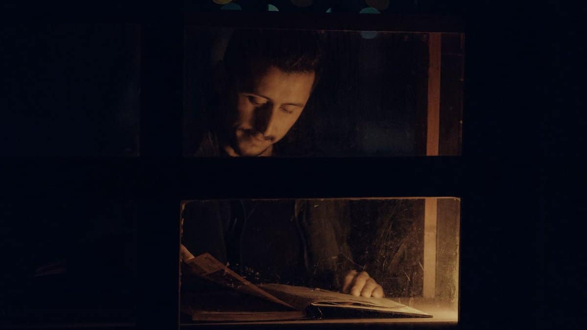 A contemplative man reading a book indoors by dim light, seen through a window at night.
