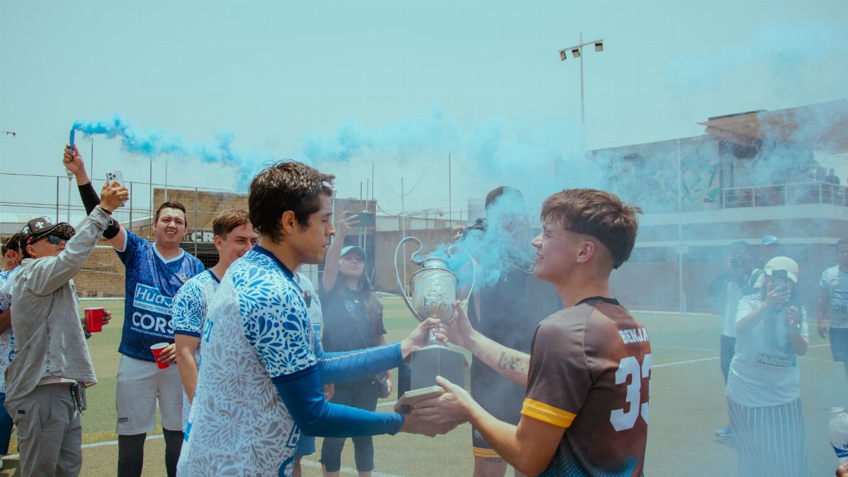 Soccer team celebrating victory with trophy, surrounded by blue smoke on outdoor field.