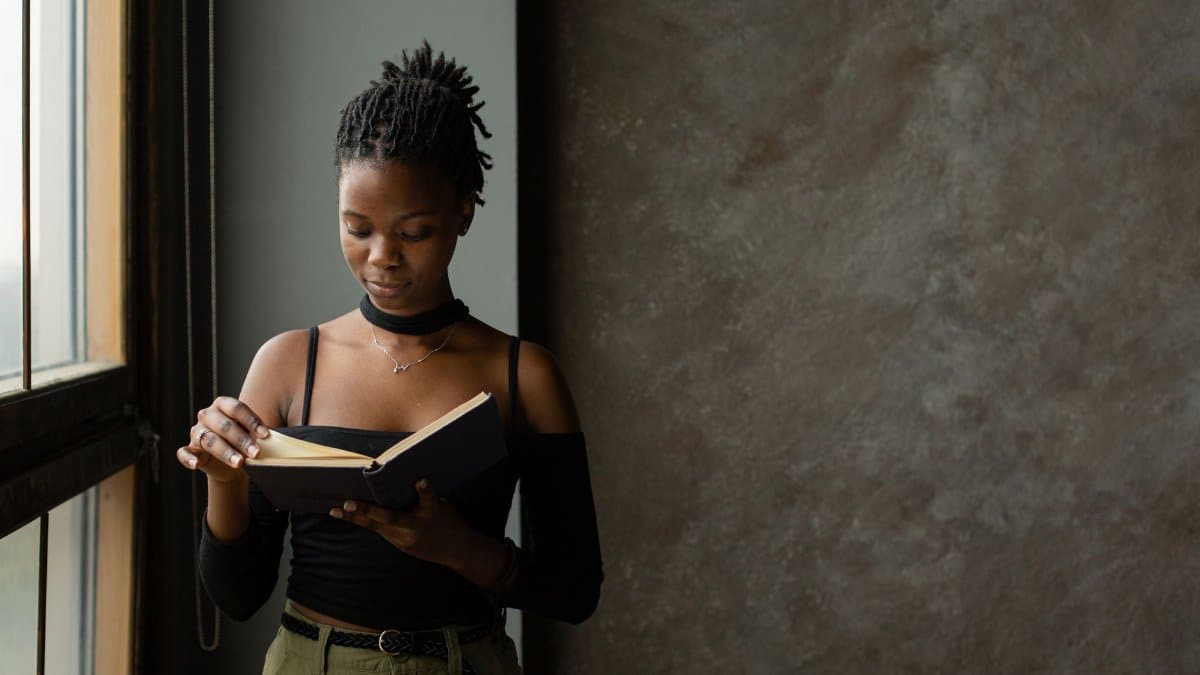 Concentrated African American female with curly dark hair in casual clothes with bare shoulders standing near window and reading book in daytime