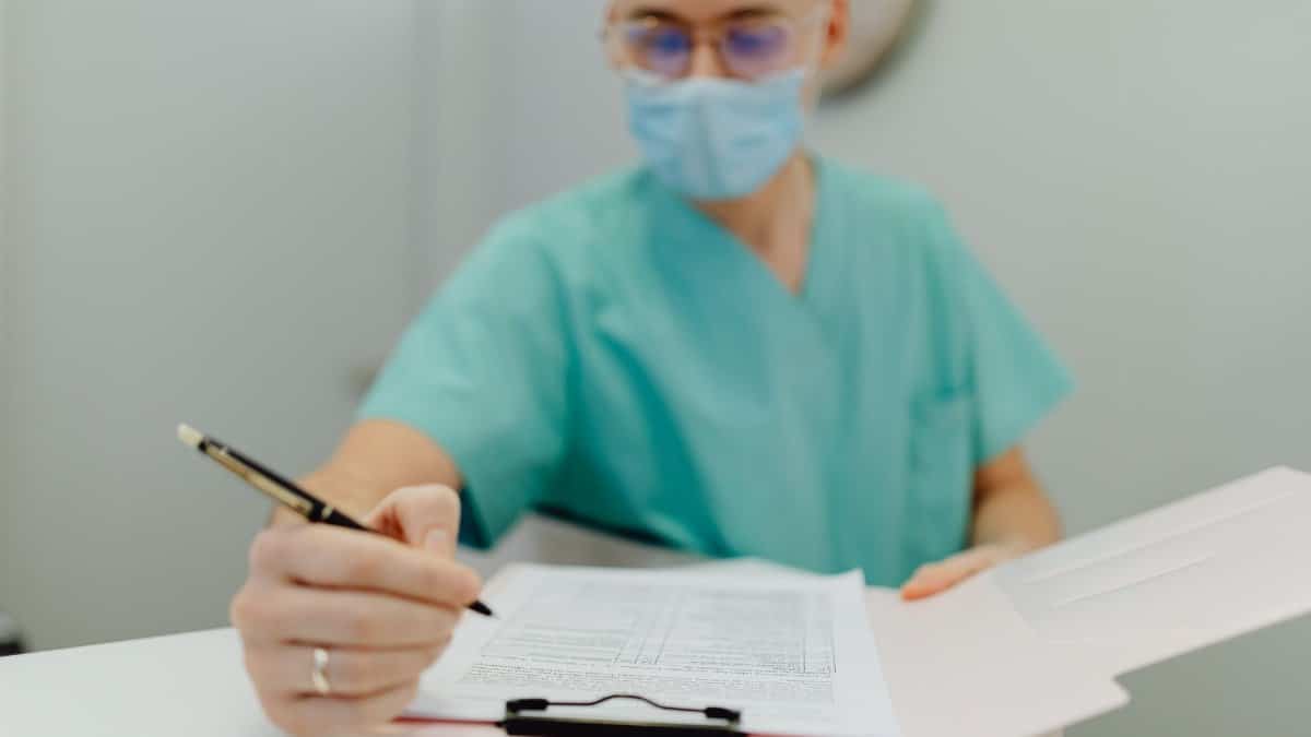 Medical practitioner wearing mask writes notes on clipboard, emphasizing patient care and safety.