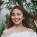 A joyful woman with braces and long hair posing outdoors in a lush garden wearing an off-shoulder top.