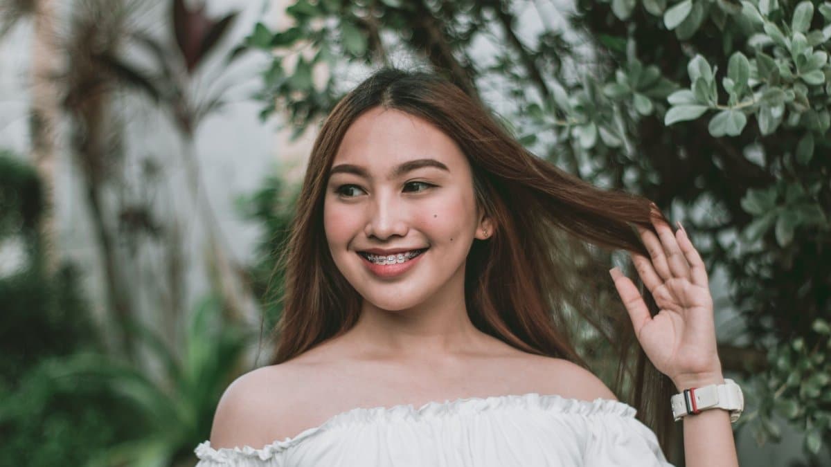 A joyful woman with braces and long hair posing outdoors in a lush garden wearing an off-shoulder top.