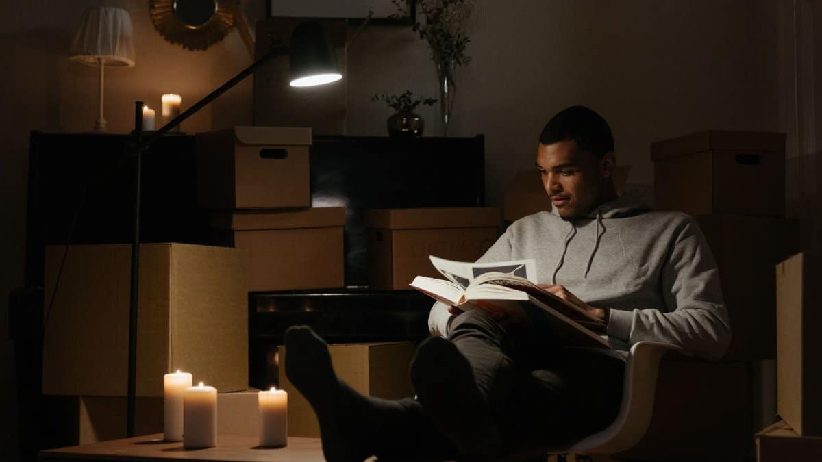 A man relaxes and reads a book in a dimly lit room filled with cardboard boxes, creating a peaceful atmosphere.