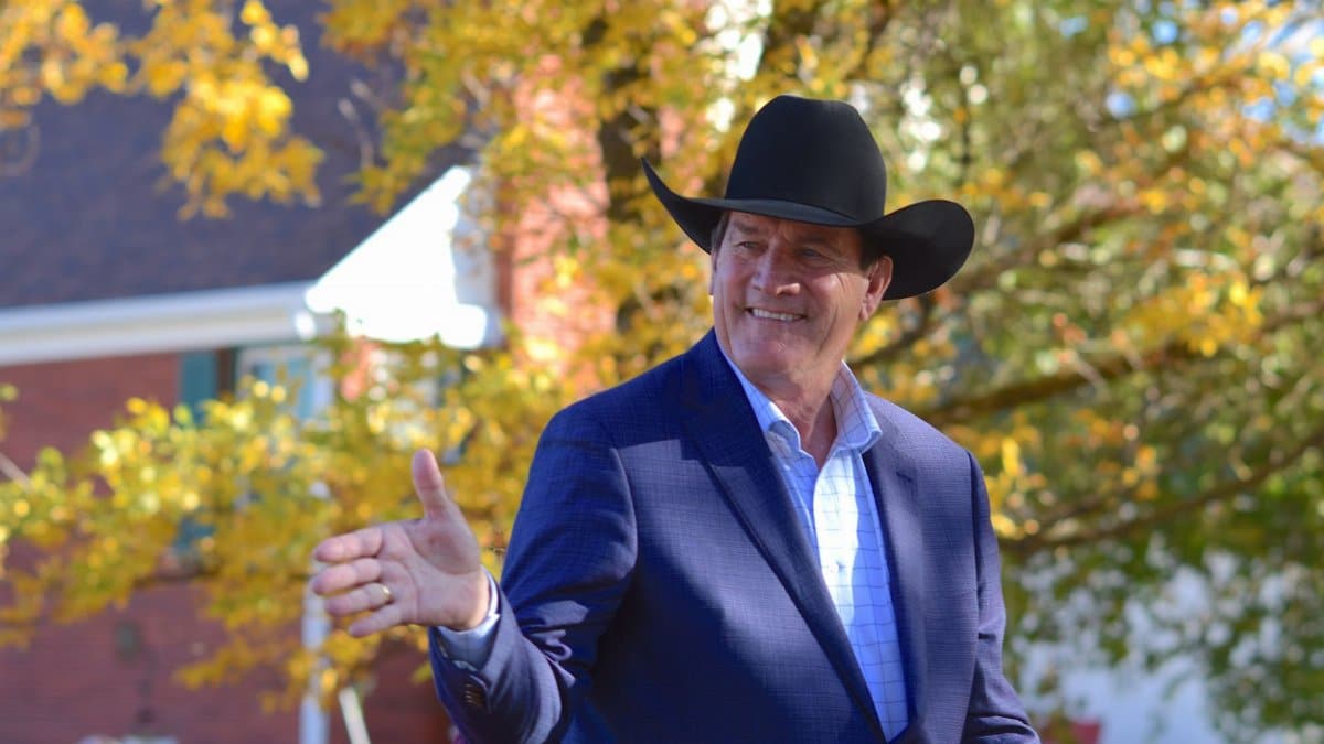 Man in a black cowboy hat and blue suit outdoors, enjoying a sunny autumn day with colorful leaves.