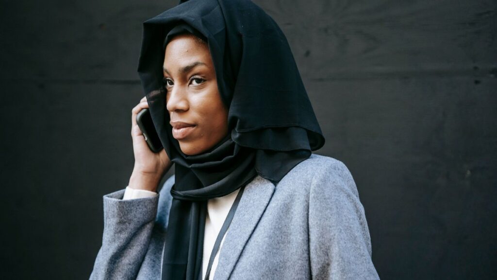 Serious young African American female in hijab and gray coat speaking on phone and looking away against black background