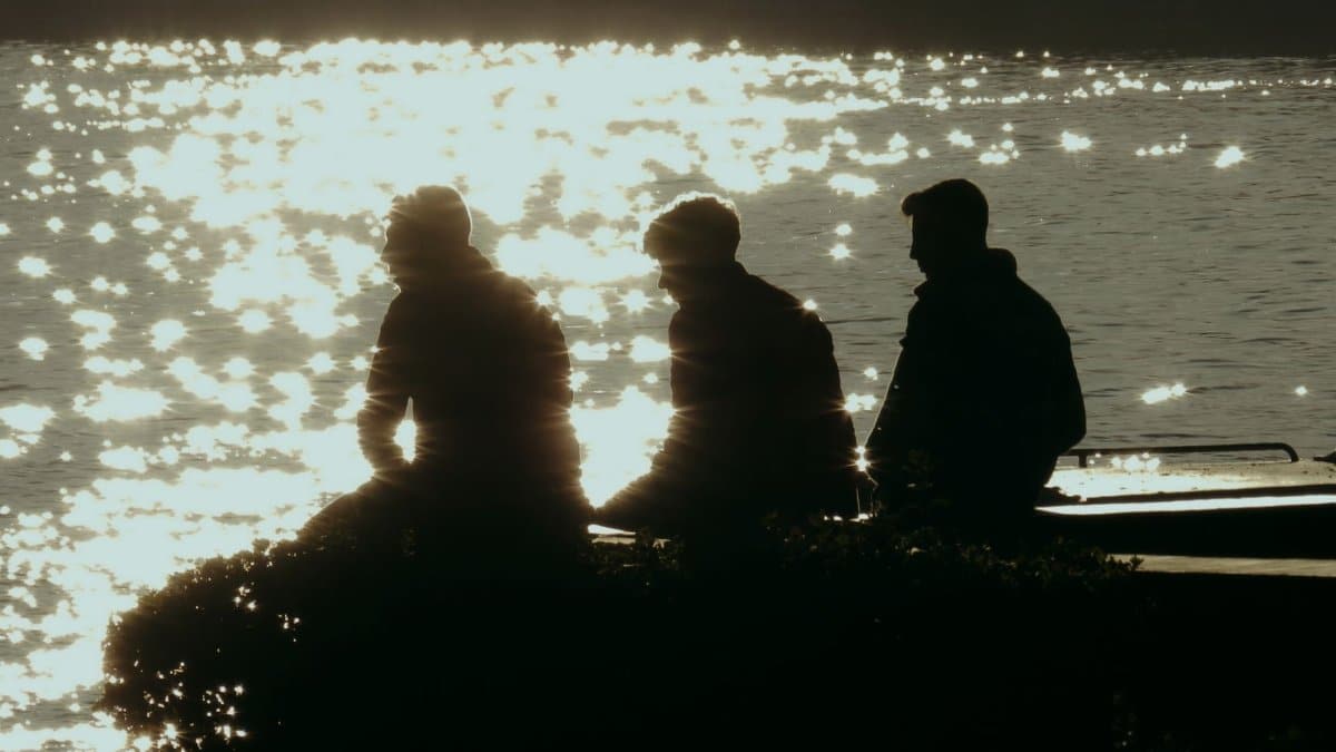 Three people silhouetted against shimmering sunlight reflecting on water, sitting by the edge.