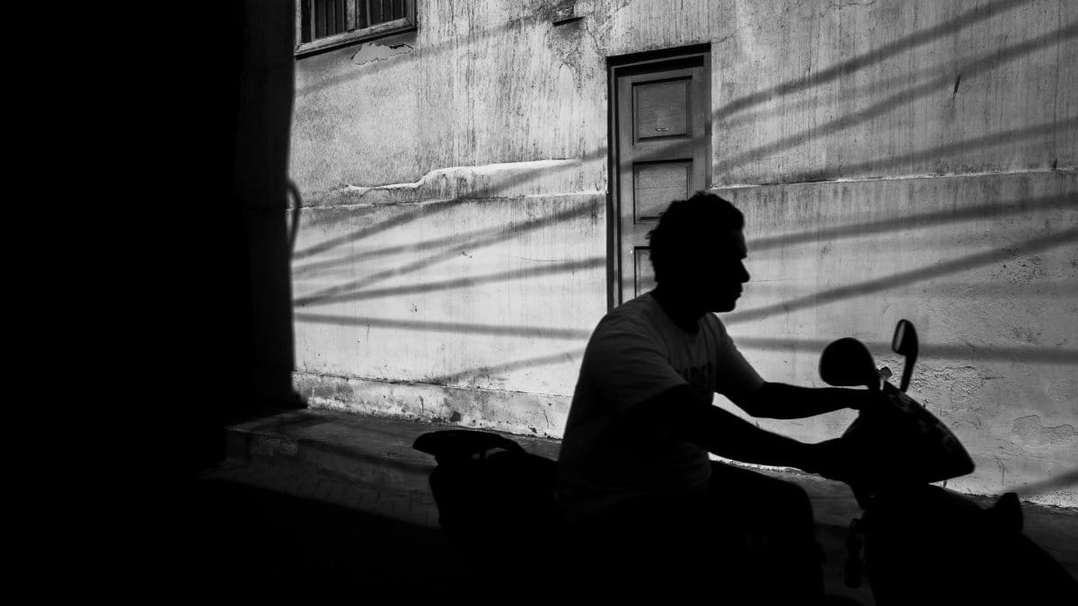 A stark black and white silhouette of a person riding a scooter against an urban wall with window shadows.