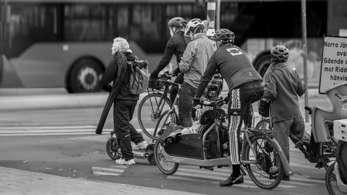 Black and white photo capturing cyclists and scooter riders waiting at a city intersection.