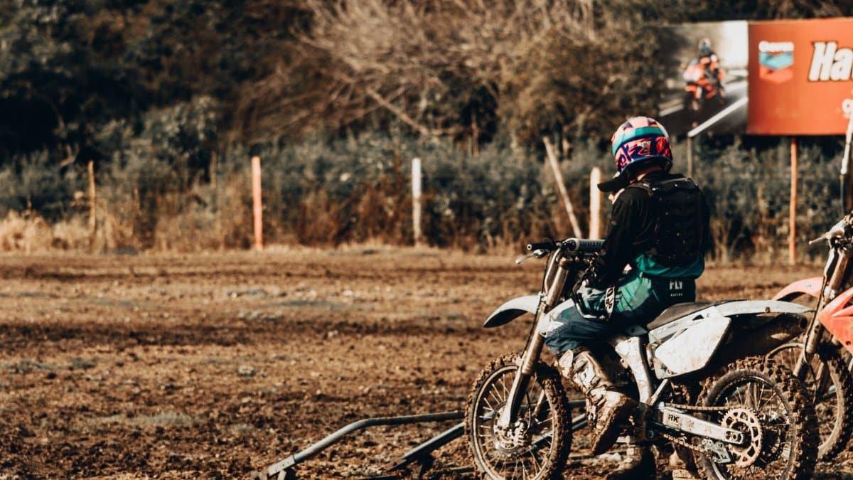 Back view of anonymous person in helmet parking motorbike on dirty ground on sunny day in countryside
