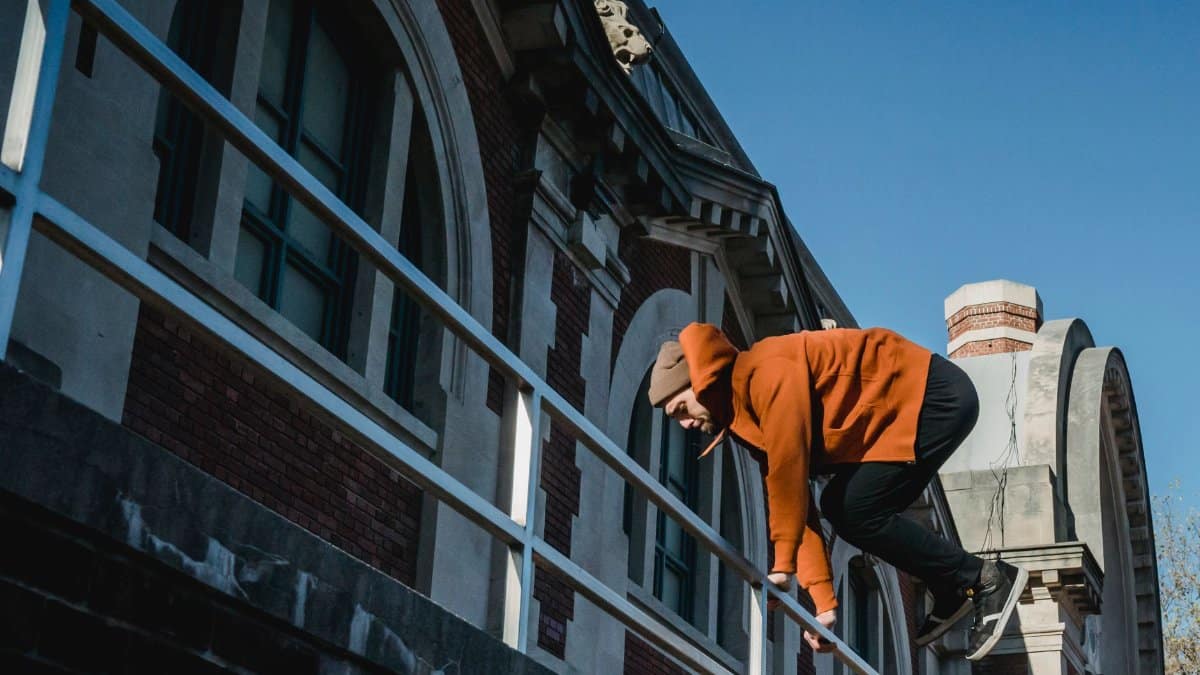 From below full length focused determined male tracer in casual hoodie jumping over brick building railing on sunny day