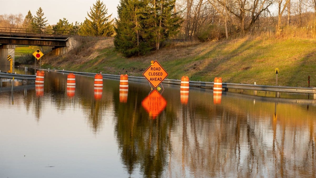 A flooded road with warning signs indicating 'Road Closed Ahead' due to water overflow.