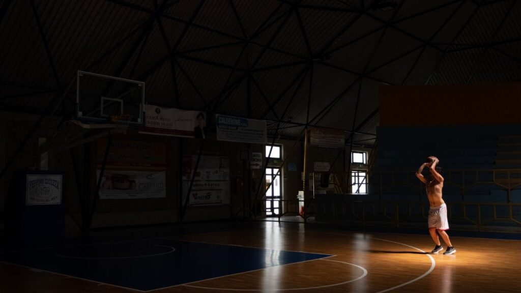 A shirtless adult male practices basketball in a dimly lit indoor court, highlighting focus and training.