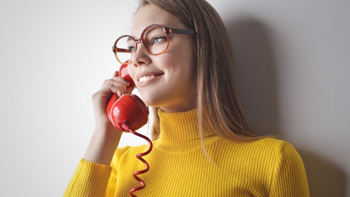 Woman in yellow sweater smiling while talking on a vintage red phone against a white wall.