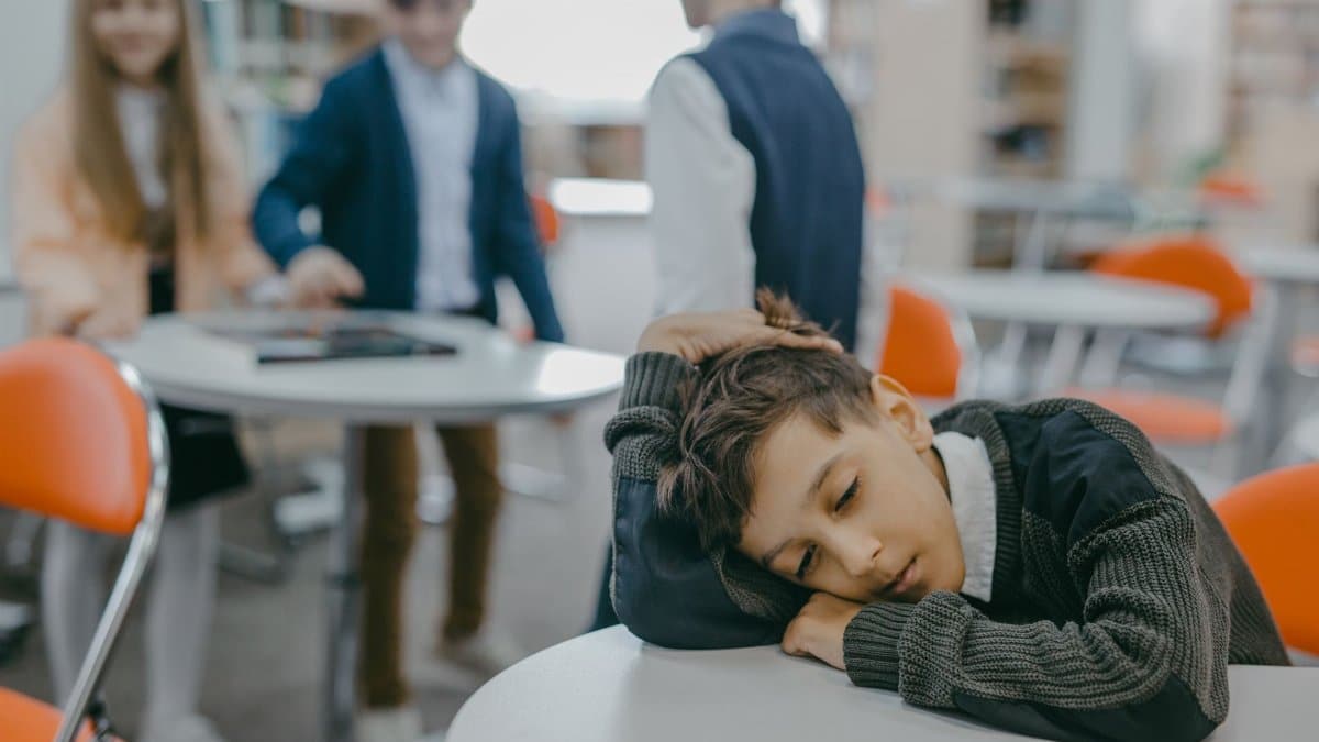 A sad child rests on a classroom desk while peers talk, symbolizing loneliness and school exclusion.