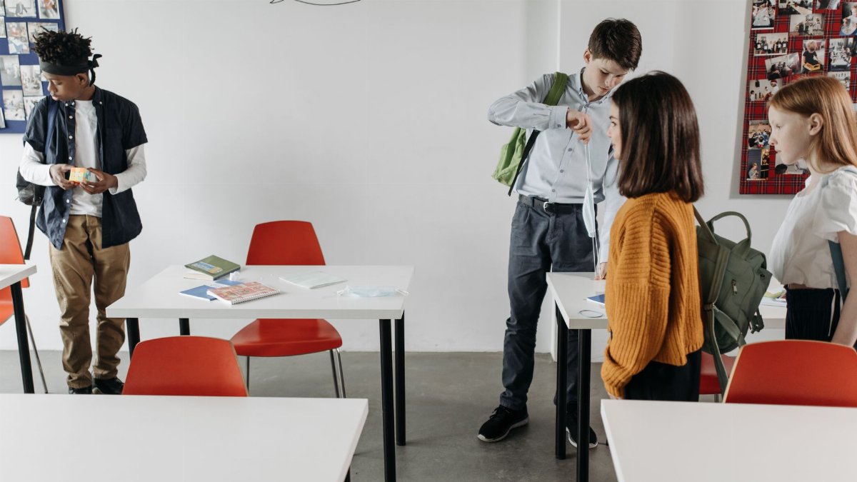 Students socializing in a classroom with red chairs and white desks, preparing to leave.