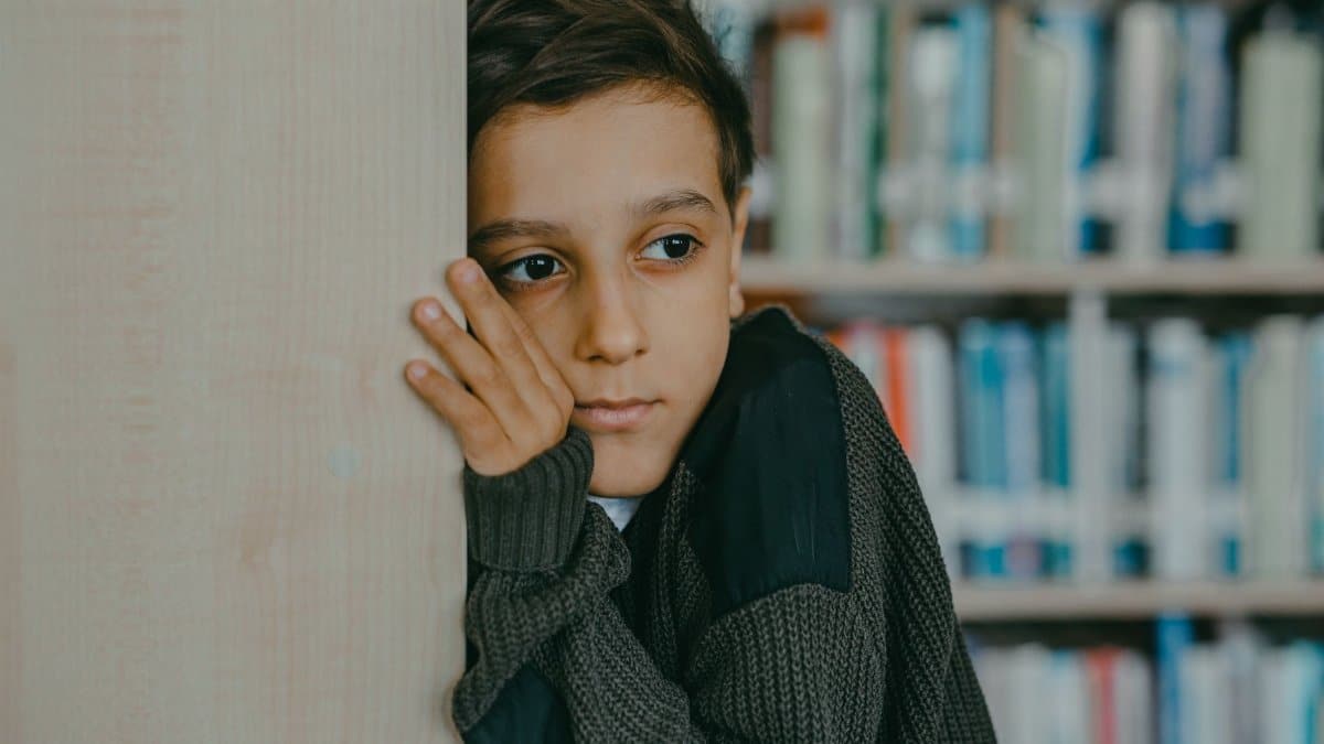A contemplative child leans against a wall in a library, deep in thought.