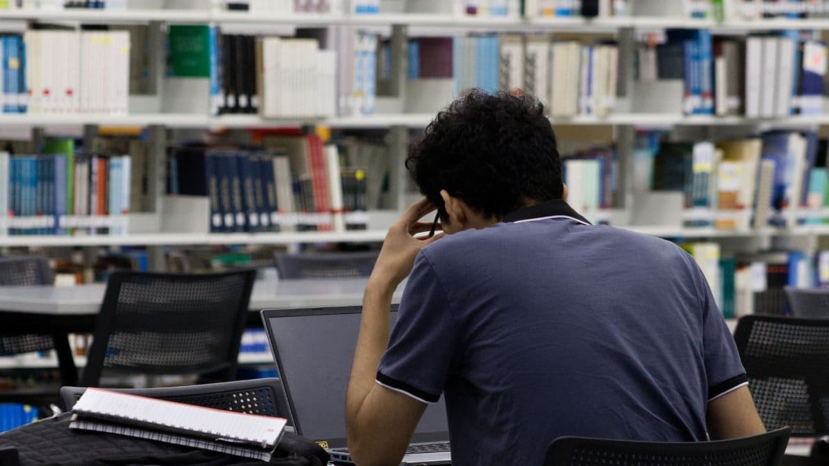 A young adult focused on studying in a Monterrey library, using a laptop and notes.