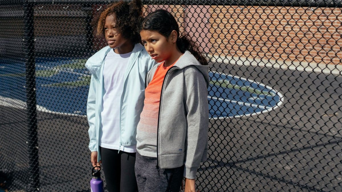 Serious multiracial girls in activewear with plastic water bottles standing near fence on sports ground during break in training