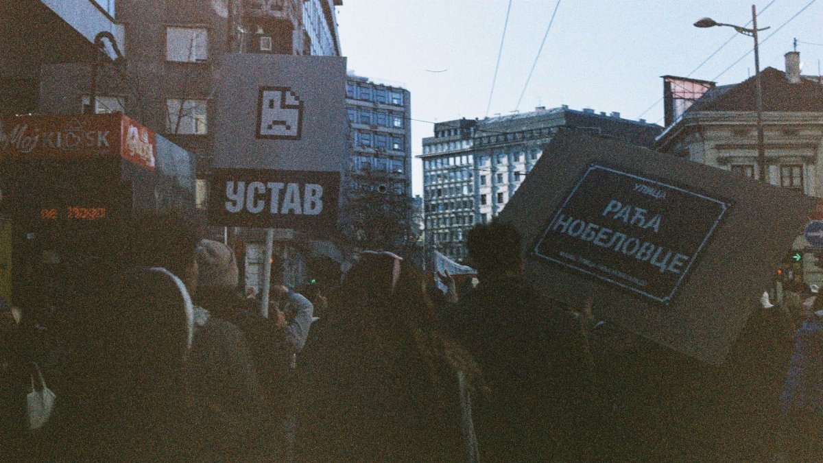 A crowd gathers for a political protest in an urban city center, holding banners and signs.