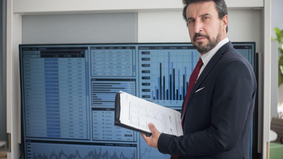 Businessman in a suit holds documents analyzing graphs on a screen, reflecting data analysis.