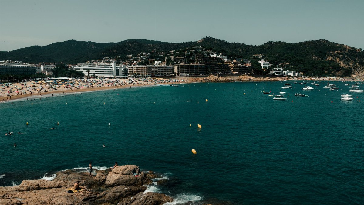 Idyllic beach setting at Tossa de Mar, Spain with crowded shoreline and boats in the bay.