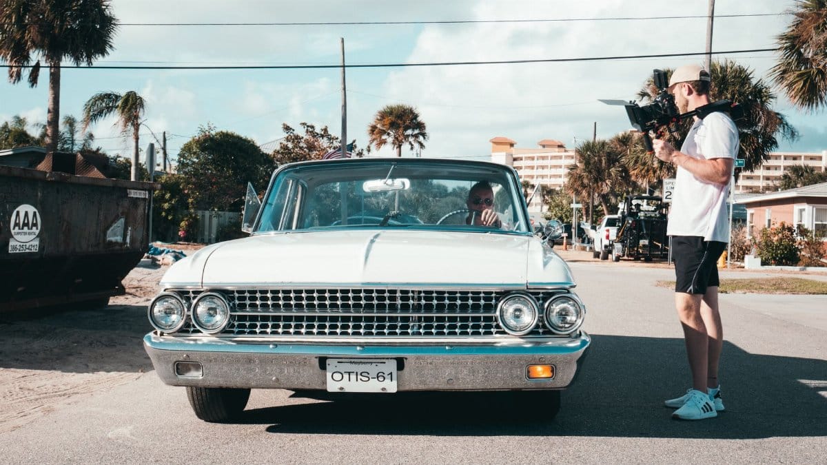 Filmmaker with handheld camera capturing a vintage car in a sunny urban street setting.