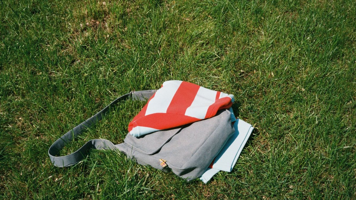 A striped blanket and grey bag rest on the grass, ready for a sunny picnic outing.