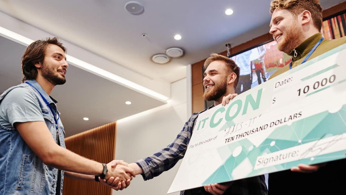 Two men celebrating with a $10,000 check at an indoor event, shaking hands and smiling.