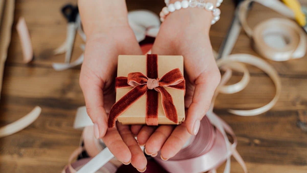 Hands presenting a small gift box wrapped with red velvet ribbon, surrounded by crafting materials.