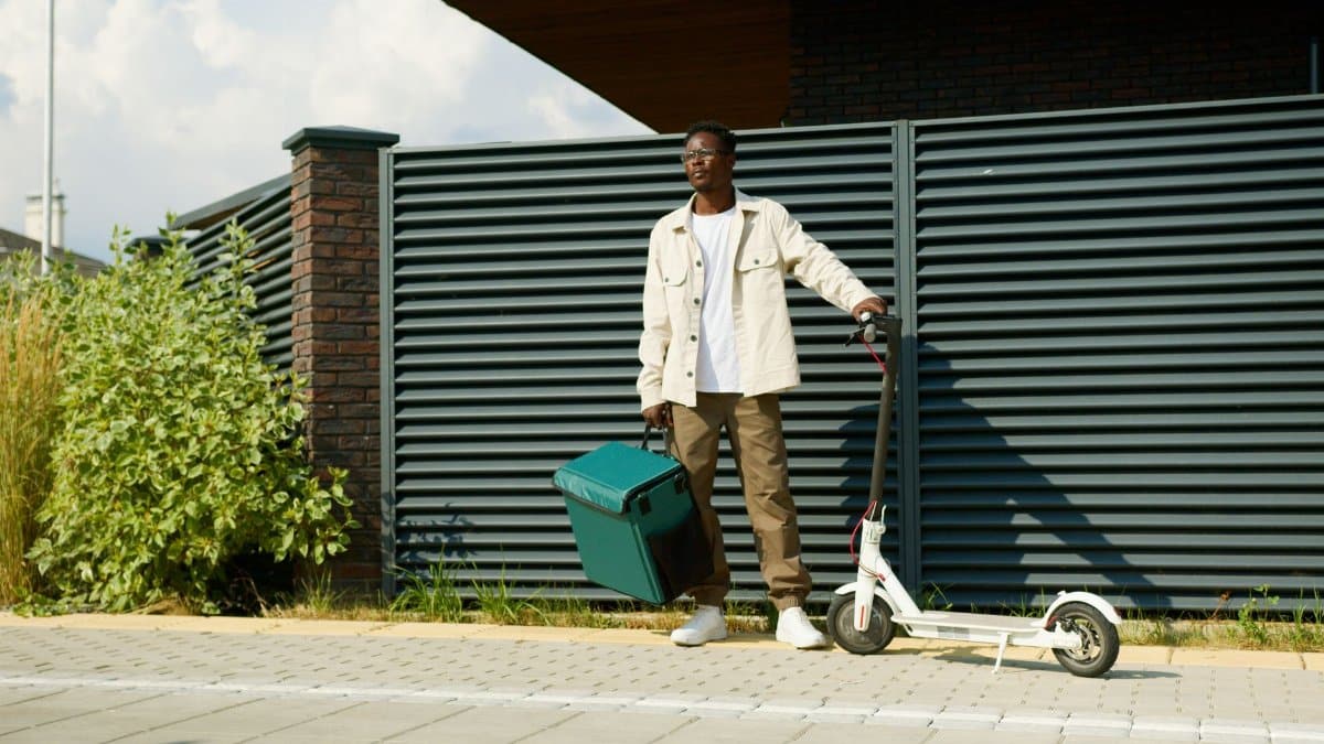 Man standing with delivery box and electric scooter outdoors.