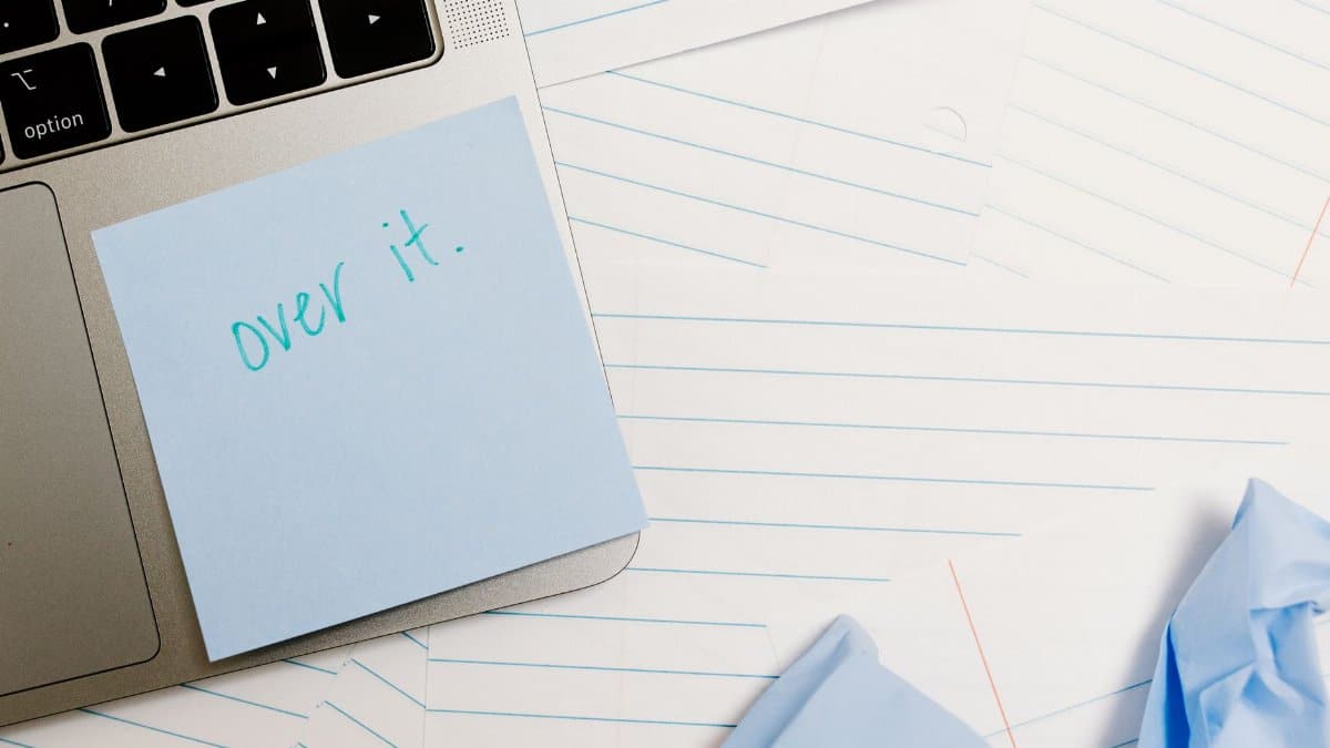 Close-up of laptop with sticky note saying 'over it' surrounded by lined papers and crumpled notes.