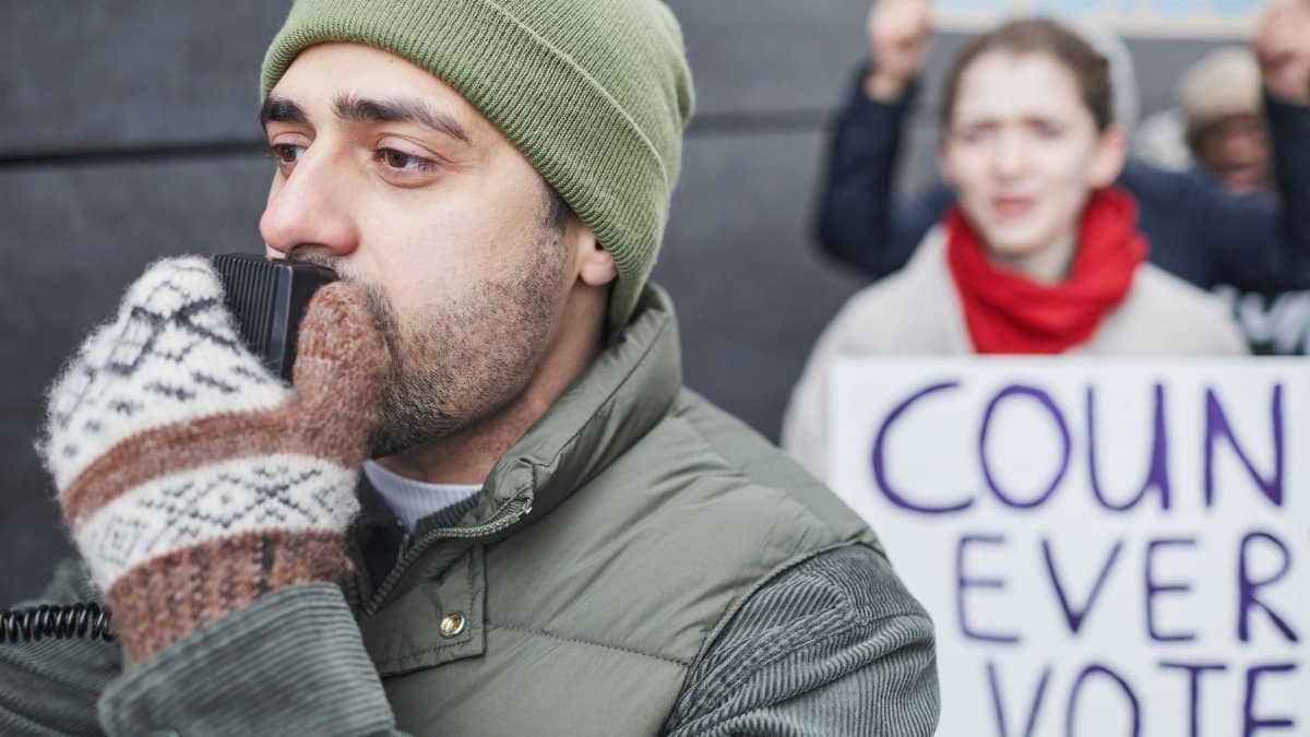A man wearing winter clothing speaks into a microphone at a protest, emphasizing the importance of voting.