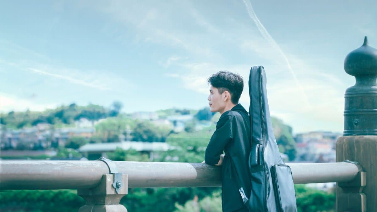 A young man with a guitar case standing on a bridge, overlooking a scenic landscape.