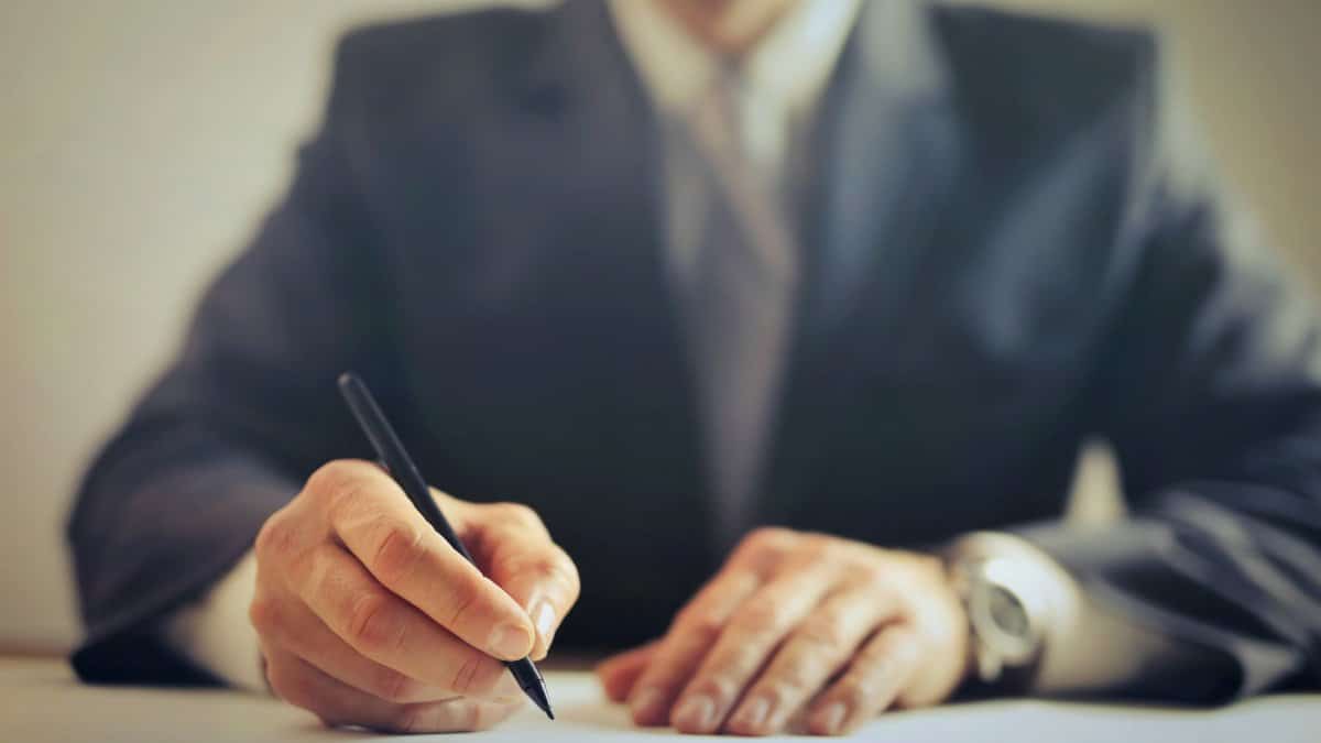 Male entrepreneur wearing formal suit sitting at table in workplace and signing documents while making successful business deal