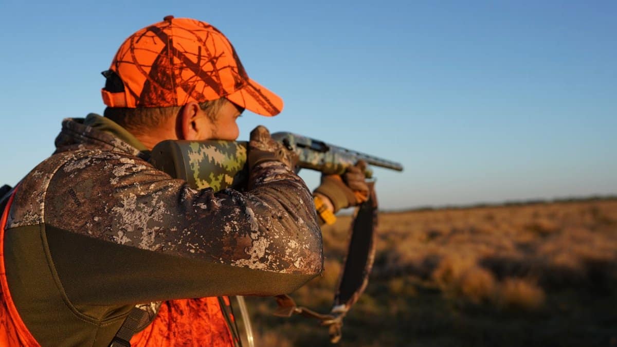 An adult man in camo and orange gear aims his rifle in a Florida field.