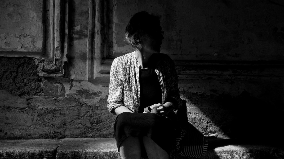 A woman sits in deep shadows against a textured wall, conveying mystery and solitude.