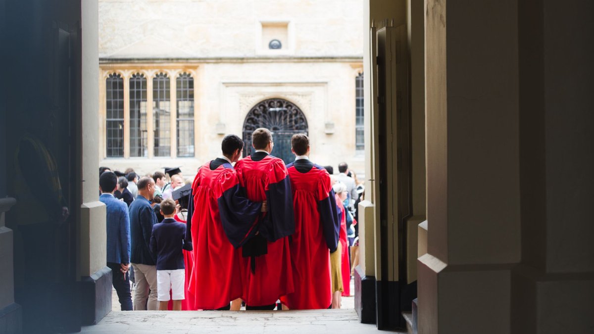 Back view unrecognizable male students wearing red cloaks bonding and standing on crowded square during graduation ceremony in historic university