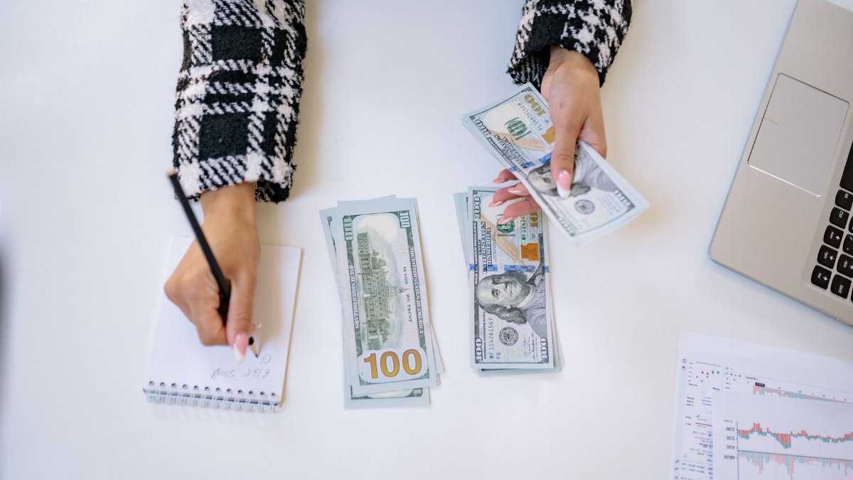 Top view of a person counting US dollars with a notepad at a workspace, indicating financial planning.