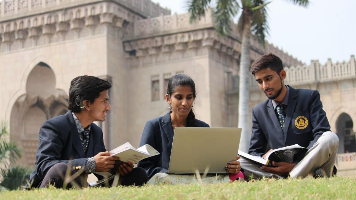 Ground level of young ethnic students in uniform sharing laptop and books while preparing for exam in yard of university