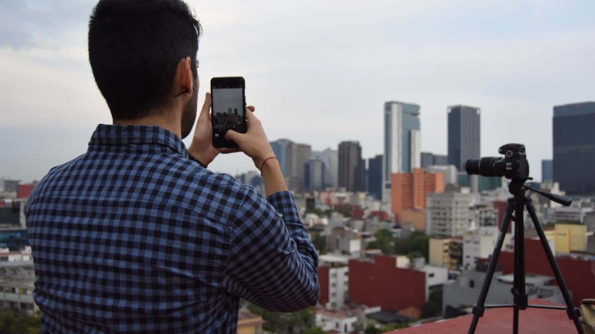 Man photographing city skyline with smartphone and camera on tripod from a rooftop.