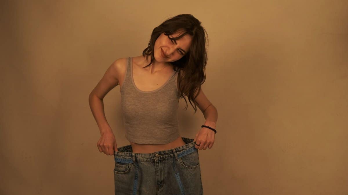 Smiling young woman in oversized jeans celebrates weight loss accomplishment.