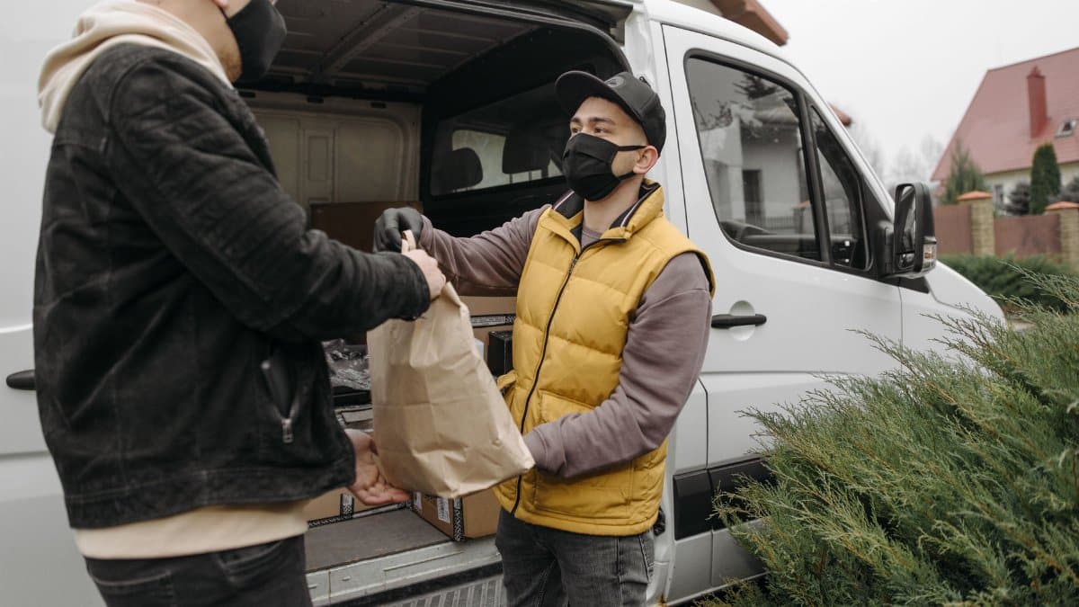 Two men wearing face masks exchange a package by a delivery van outdoors.